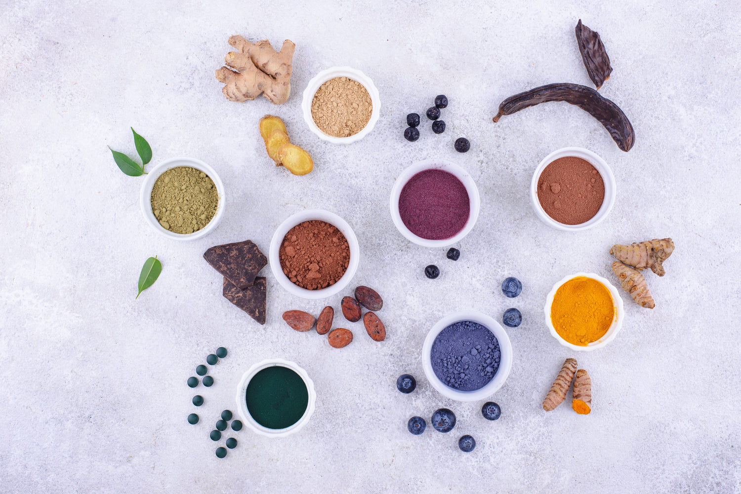 Assorted superfood powders and natural ingredients, including turmeric, cacao, spirulina, ginger, and blueberries, displayed on a white background